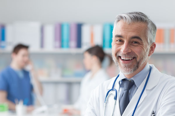 médico sonriente mirando a cámara, con dos compañeros de fondo en la consulta
